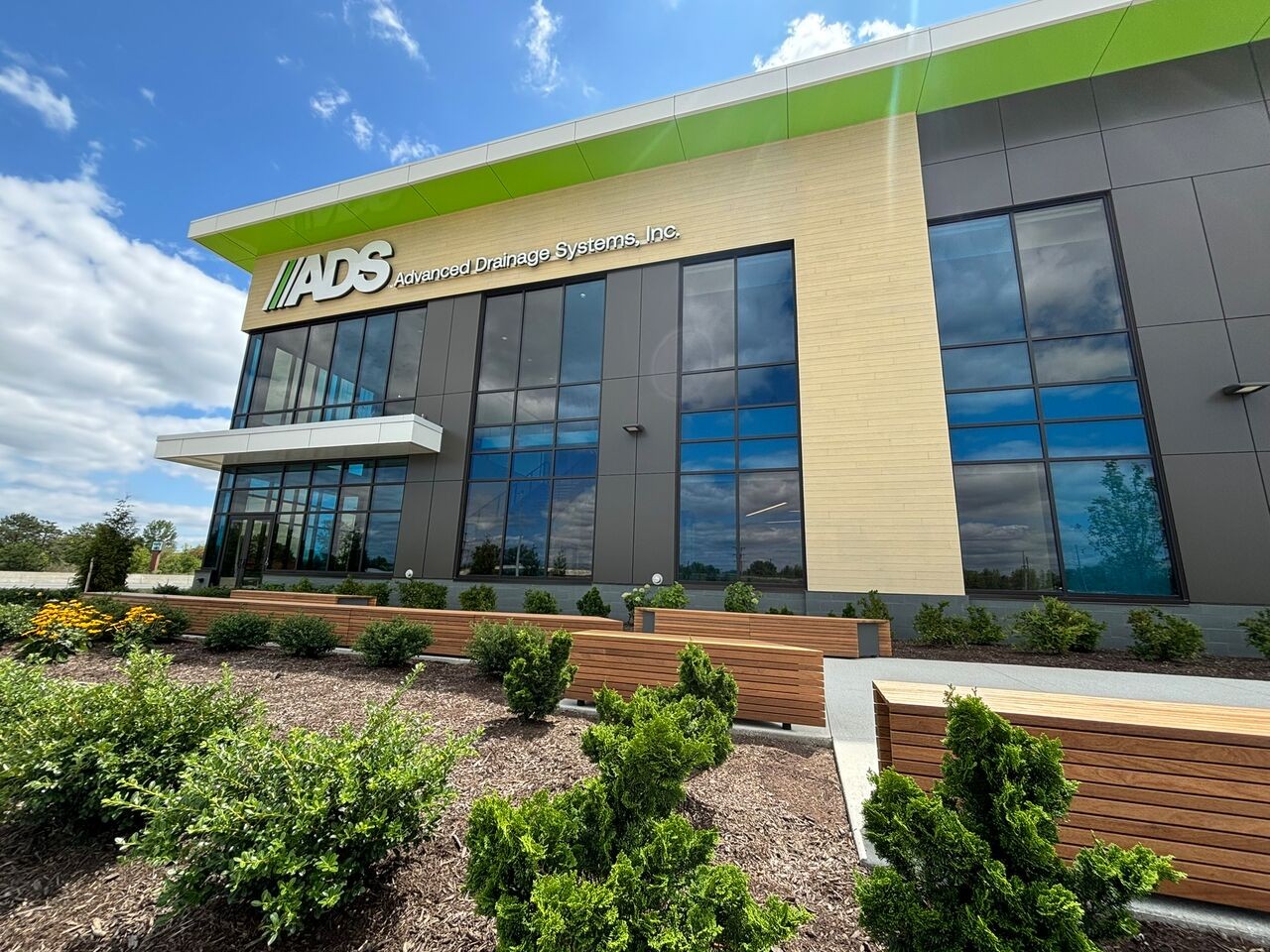 Exterior view of the Advanced Drainage Systems Engineering and Technology Center building in Hilliard, Ohio, featuring modern glass windows, light tan facade panels, green roof trim, and landscaped ground with wooden benches.
