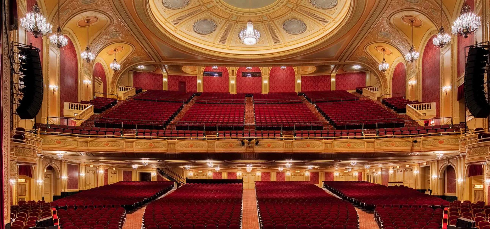 Symmetrical interior view of the Palace Theatre auditorium showing red plush seating, gilded balcony, and illuminated domed ceiling.