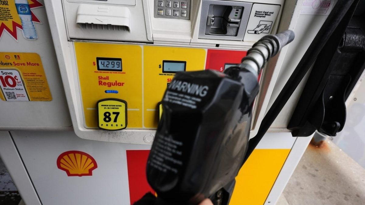 Close-up of a fuel nozzle being lifted at a Shell gas station pump showing a price of $4.299 per gallon for regular 87 octane gasoline.