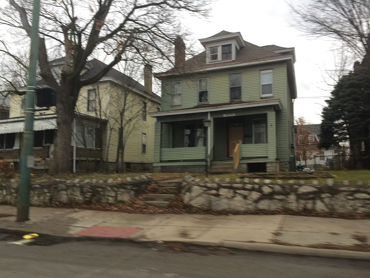 Two-story residential homes in the Hilltop neighborhood of Columbus, Ohio, with a green siding house and stone retaining wall along the sidewalk.