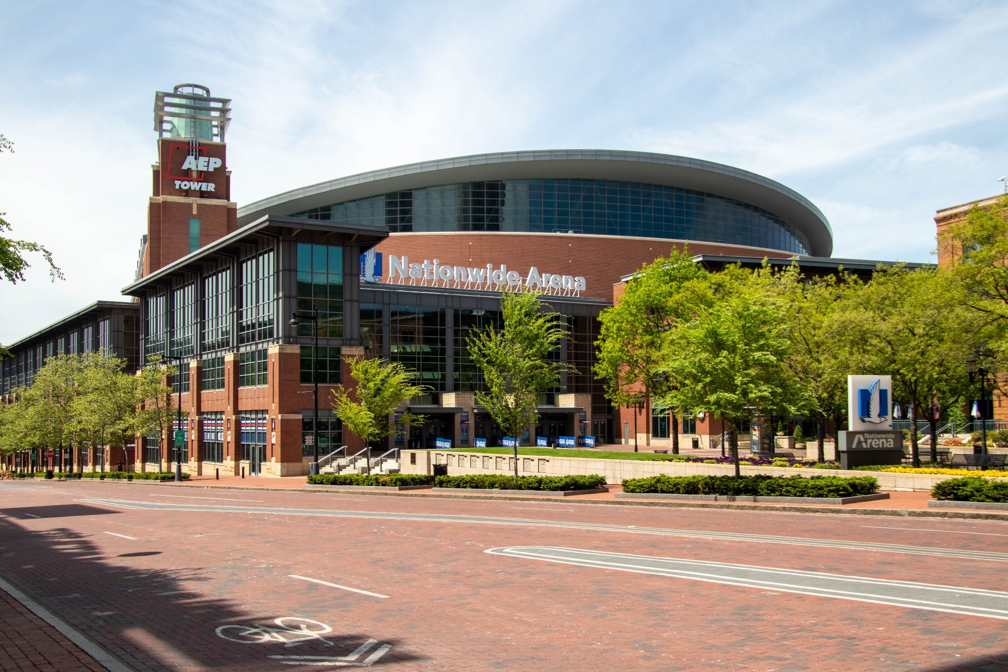 Daytime exterior view of Nationwide Arena featuring its brick facade, glass entrance, and curved roofline along a tree-lined street.