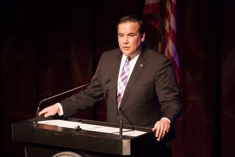 Columbus Mayor Andrew Ginther speaking at a lectern with two microphones and an American flag in the background.