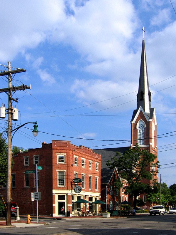 A historic brick street corner in German Village, Columbus, with street trees, an outdoor cafe, and a tall church steeple.