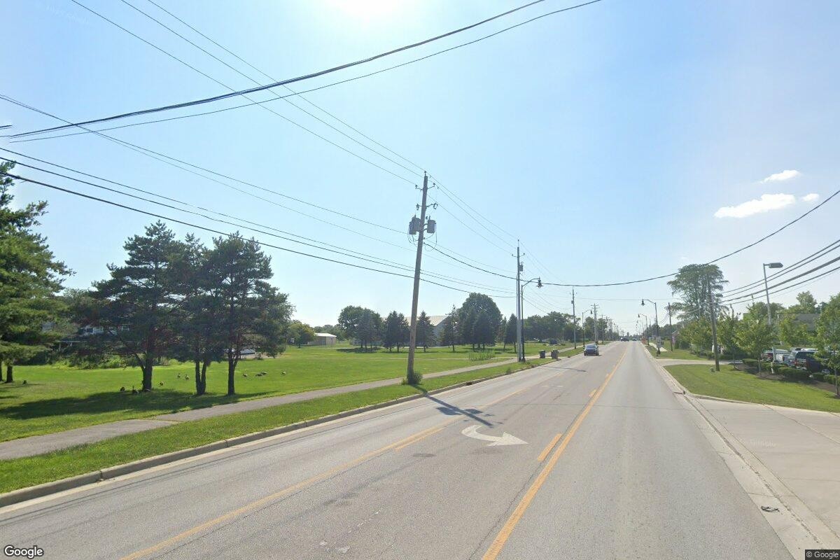 Paved two-lane road flanked by open green fields and trees on the left and commercial curbside development on the right.