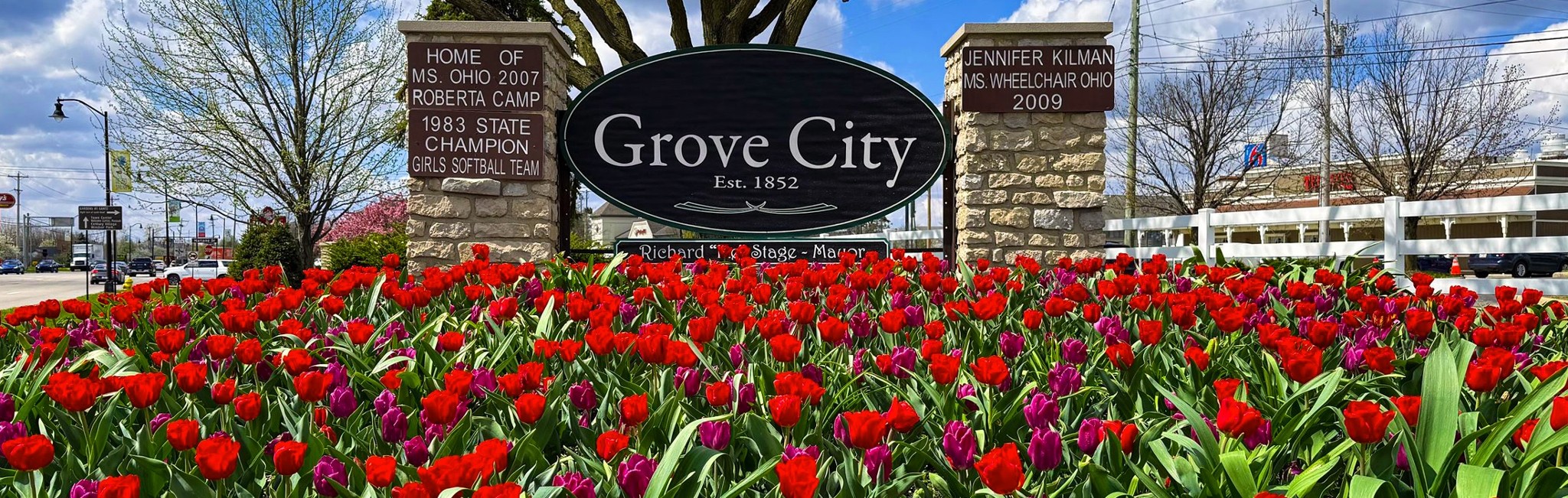 A prominent stone and oval wooden welcome sign reading Grove City Est. 1852, flanked by stone pillars and set behind a dense garden bed of blooming red and purple tulips.