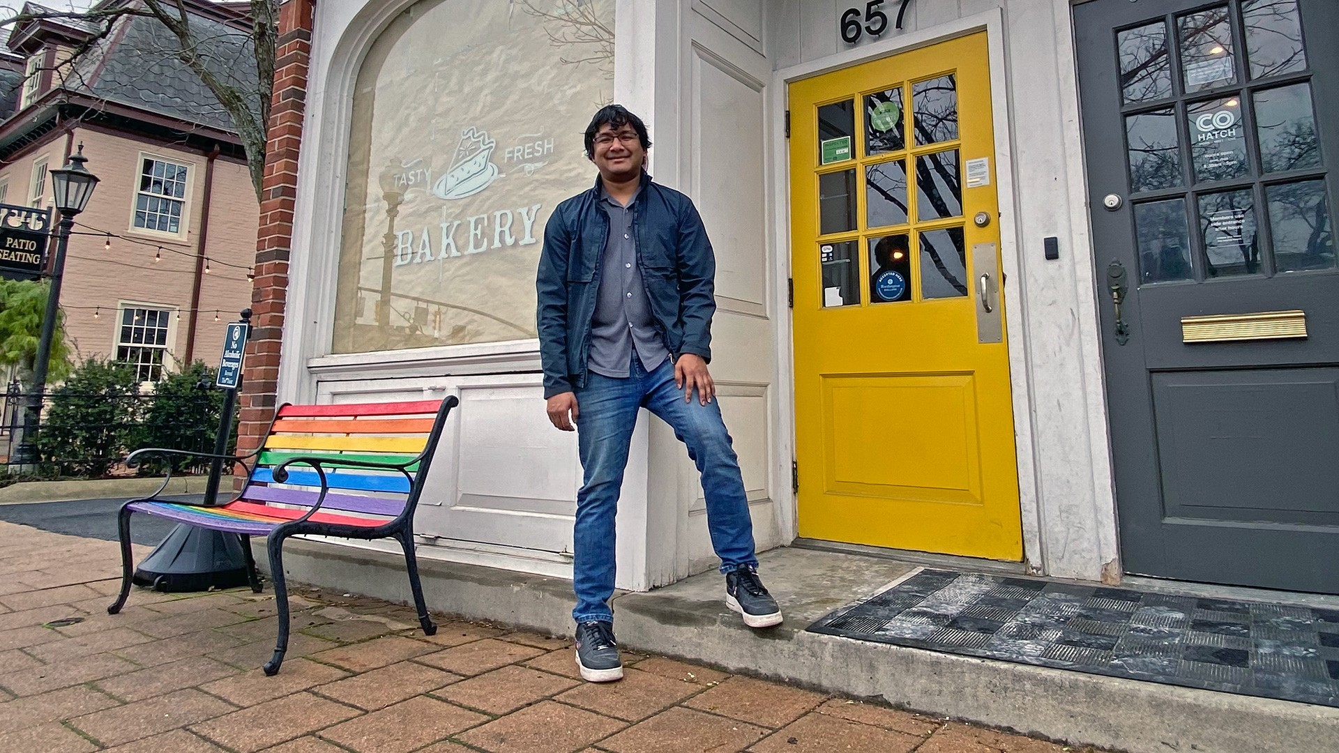 Chef Avishar Barua standing outside the storefront entrance of Joya's cafe next to a bright yellow door and rainbow bench in Old Worthington.