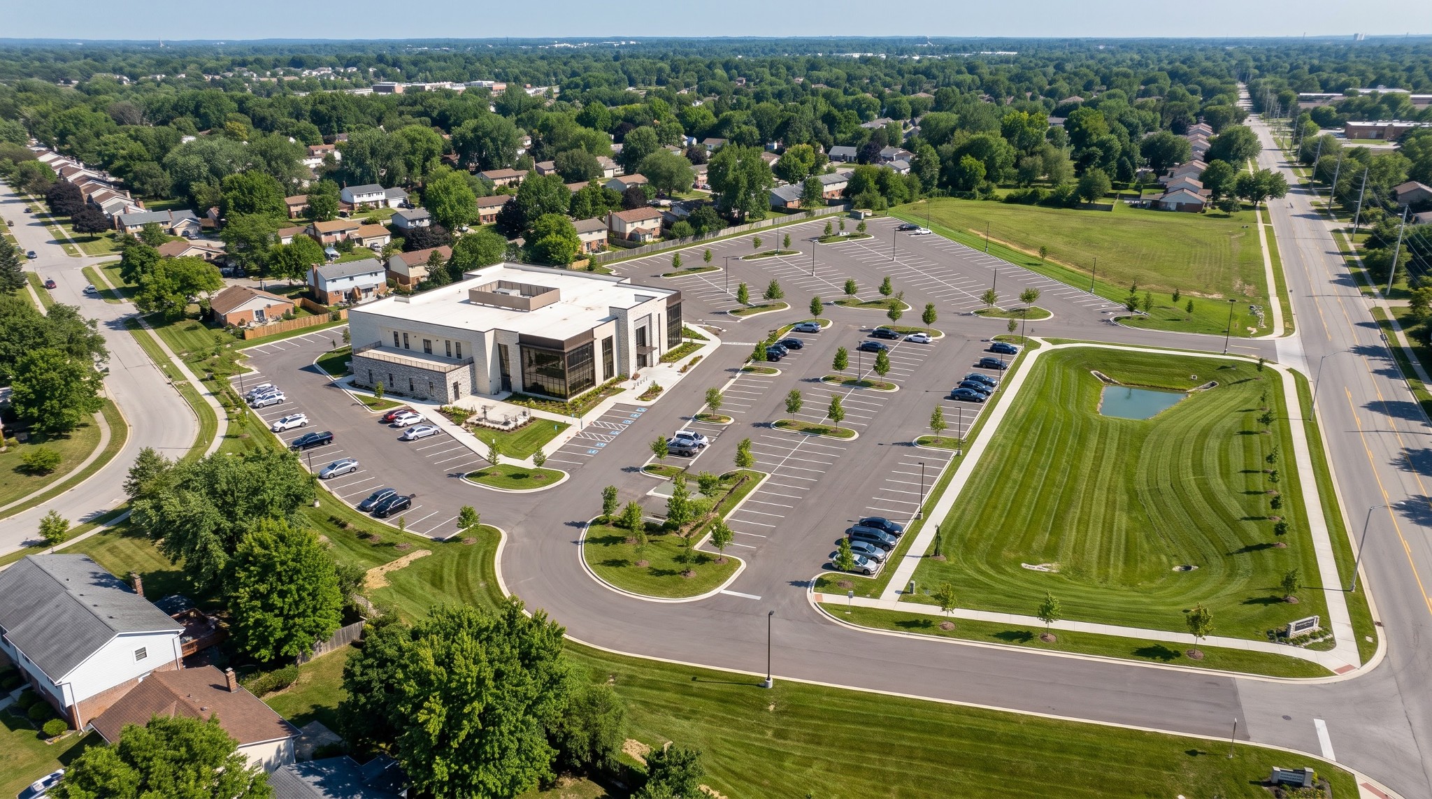 Conceptual overhead rendering of a modern two-story building, large surface parking lot, and stormwater detention basin in a suburban setting.