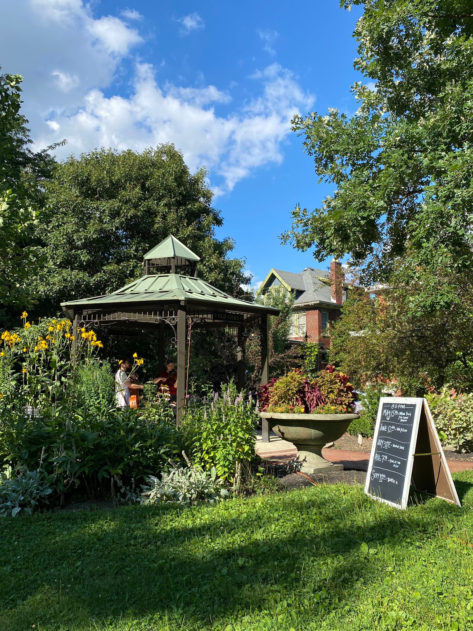 A green metal gazebo marked 'Old Oaks' surrounded by flowering garden beds and mature trees, with musicians performing inside and a chalkboard concert schedule in the foreground.