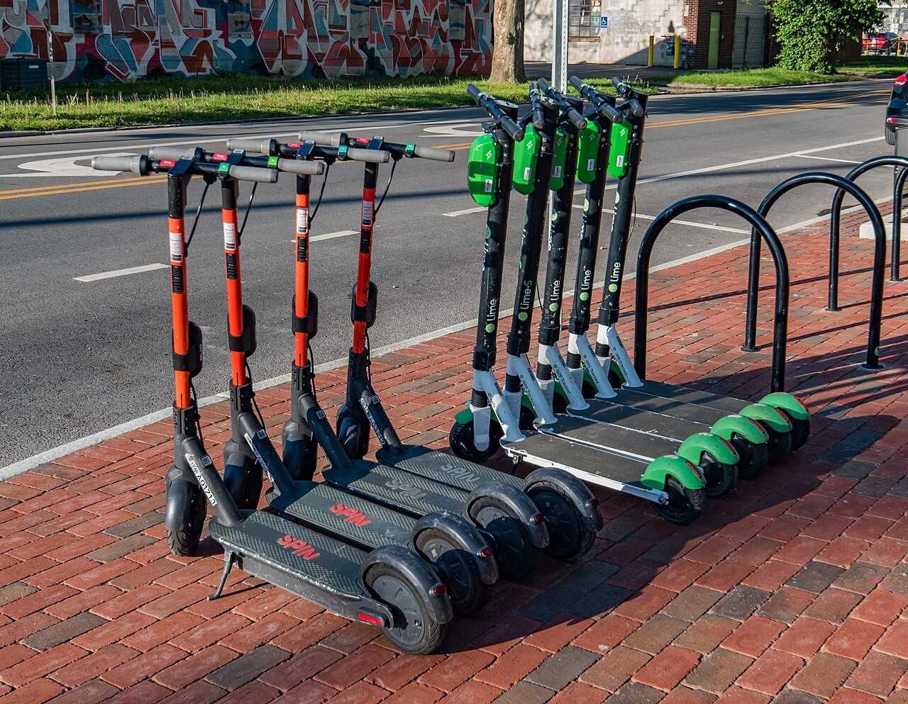 Seven electric scooters from Spin and Lime parked in a row on a red brick sidewalk beside a roadway in Columbus, Ohio.