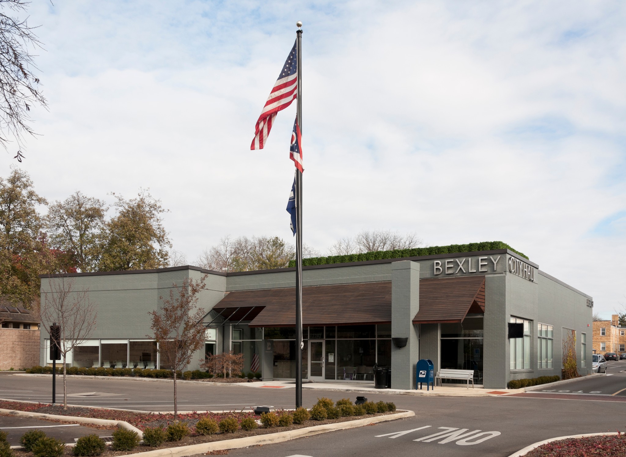 Exterior view of Bexley City Hall, a modern grey brick municipal building with glass windows, brown canopies, and a tall flagpole displaying the American, Ohio, and Bexley flags.