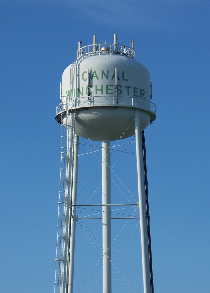A tall white municipal water tower with green lettering reading Canal Winchester against a clear blue sky.