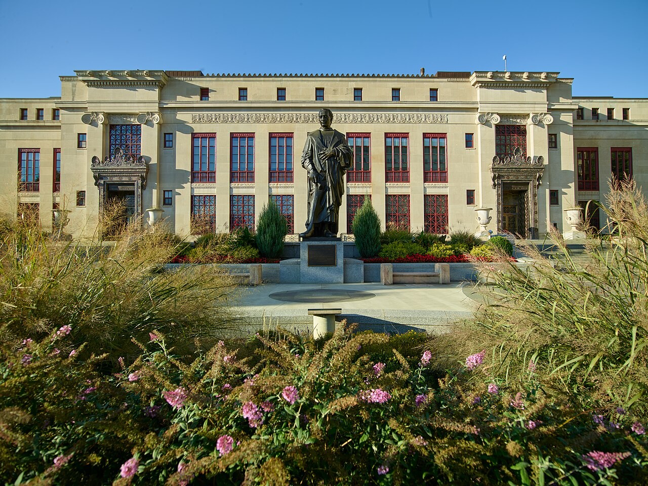 Bronze statue of Christopher Columbus on a pedestal in front of Columbus City Hall's neoclassical limestone facade, framed by flowering shrubs and ornamental grasses.