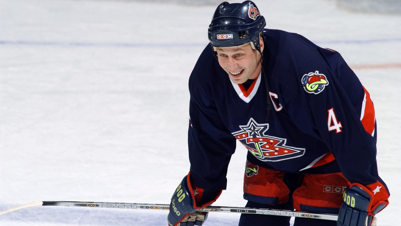 Defenseman Lyle Odelein smiles while resting on his hockey stick on the ice in a Columbus Blue Jackets dark blue jersey with captain's C.