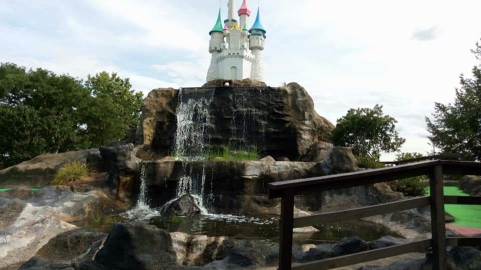 A miniature white fairytale castle with colorful spires perched above a tiered stone waterfall alongside a miniature golf putting green.