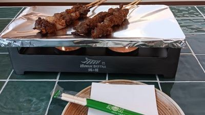 Grilled skewers on a branded foil-lined tabletop warmer with chopsticks on a green tiled table.