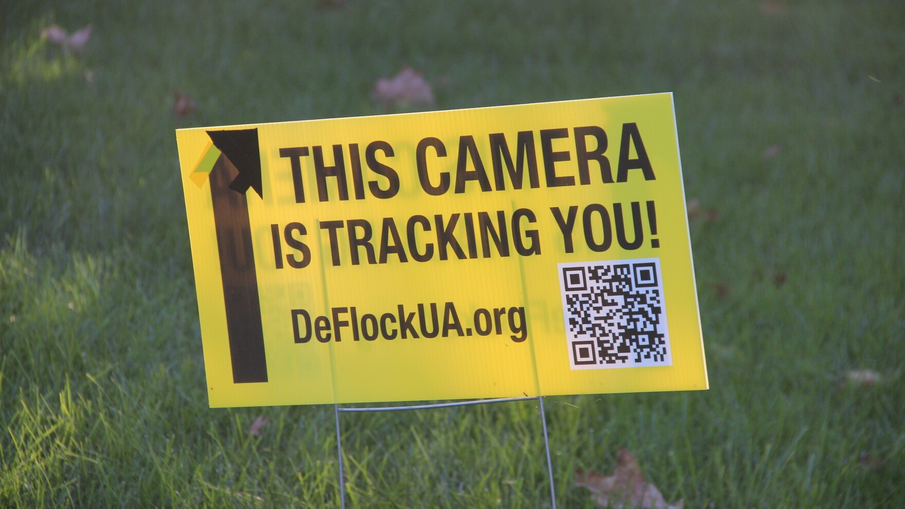 A yellow lawn sign reading 'THIS CAMERA IS TRACKING YOU!' with an arrow and DeFlockUA.org website.