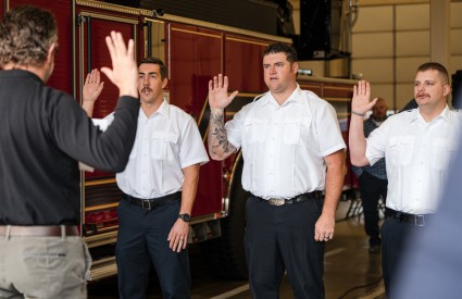 Three Upper Arlington firefighters in white uniform shirts raising their right hands taking the oath of office in front of a red fire engine.