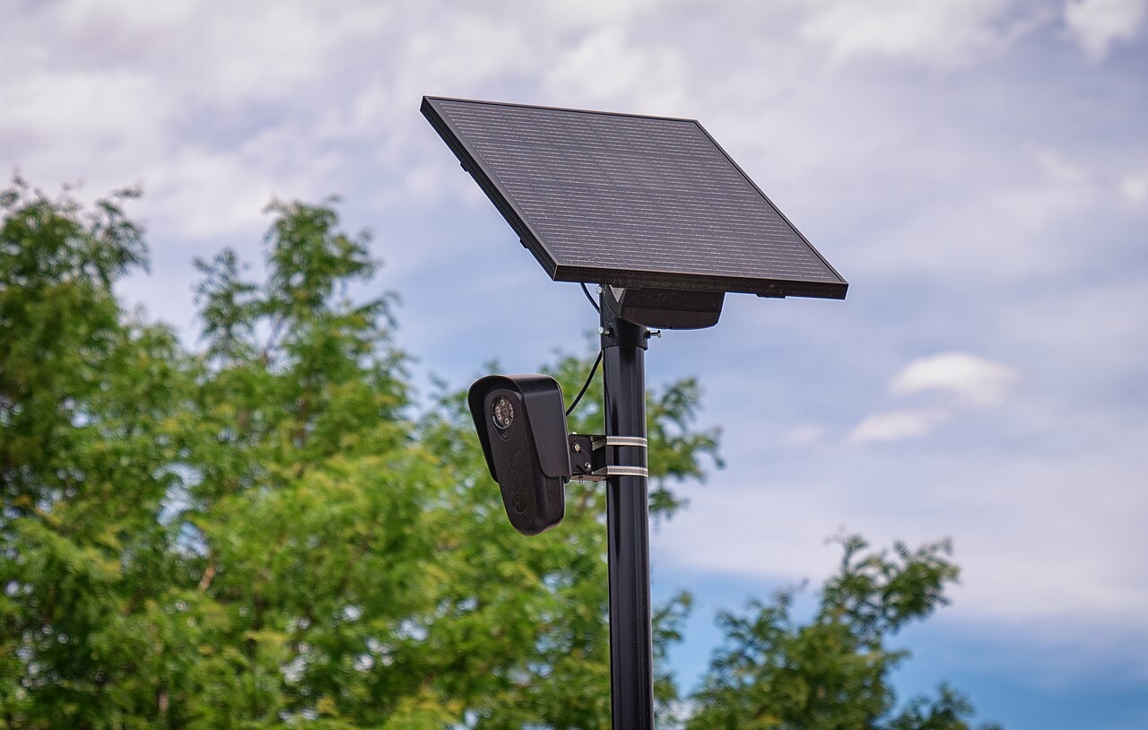 A Flock Safety automated license plate reader camera and solar panel mounted on a black pole against foliage and cloudy sky.