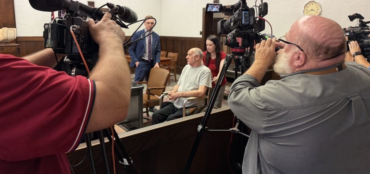 Gary Siders Sr. seated in a wheelchair in a wood-paneled courtroom with attorney and camera crew present.