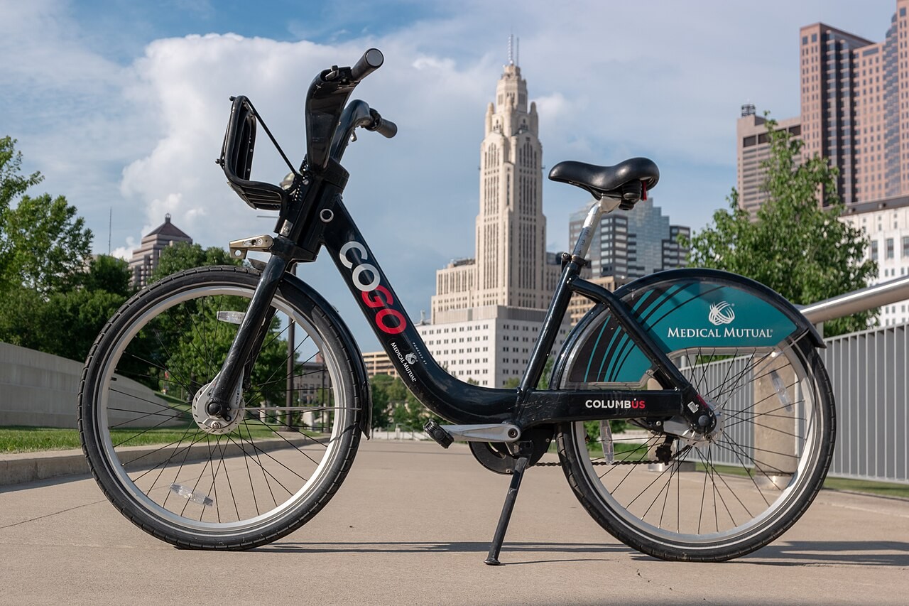 A black CoGo bike share bicycle parked on a paved promenade with downtown Columbus buildings in the background.