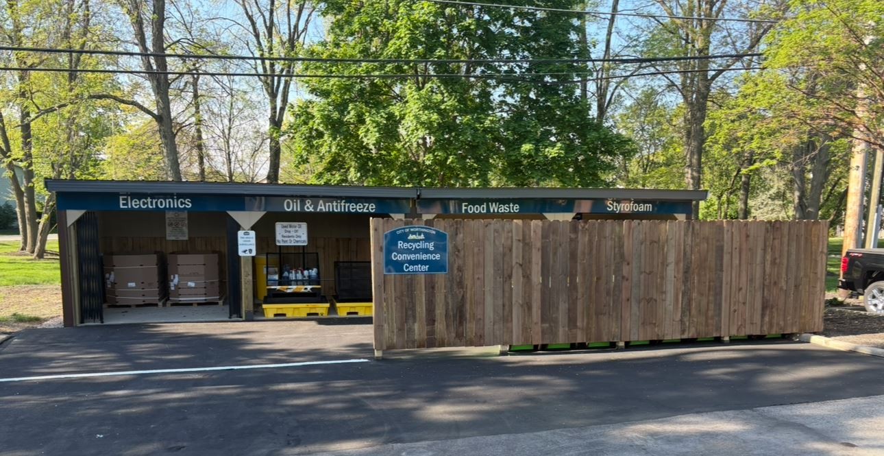 Covered collection bays at the City of Worthington Recycling Convenience Center labeled Electronics, Oil and Antifreeze, Food Waste, and Styrofoam behind a wooden fence with an official city sign.